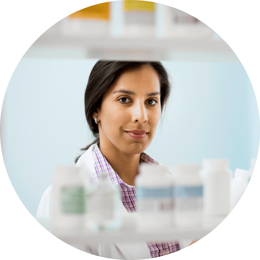 A woman in a lab coat stands in front of a shelf of bottles, representing a laboratory setting and scientific work.